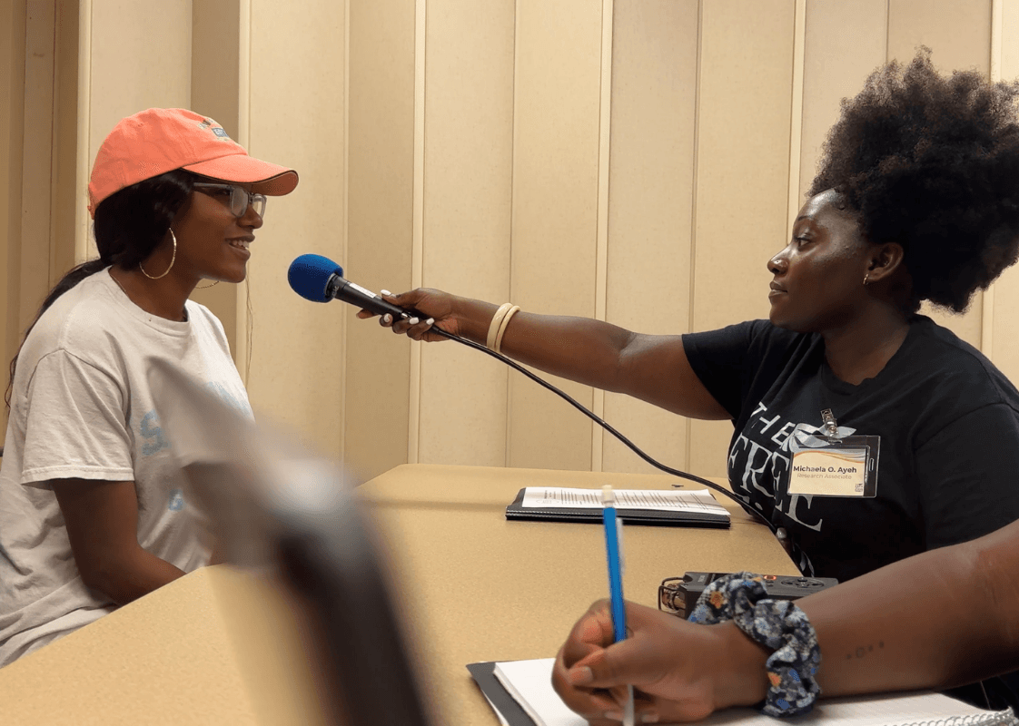A female student wearing a black T-shirt holds a microphone up to a woman wearing a baseball cap for an interview; both sit across from one another at a table.